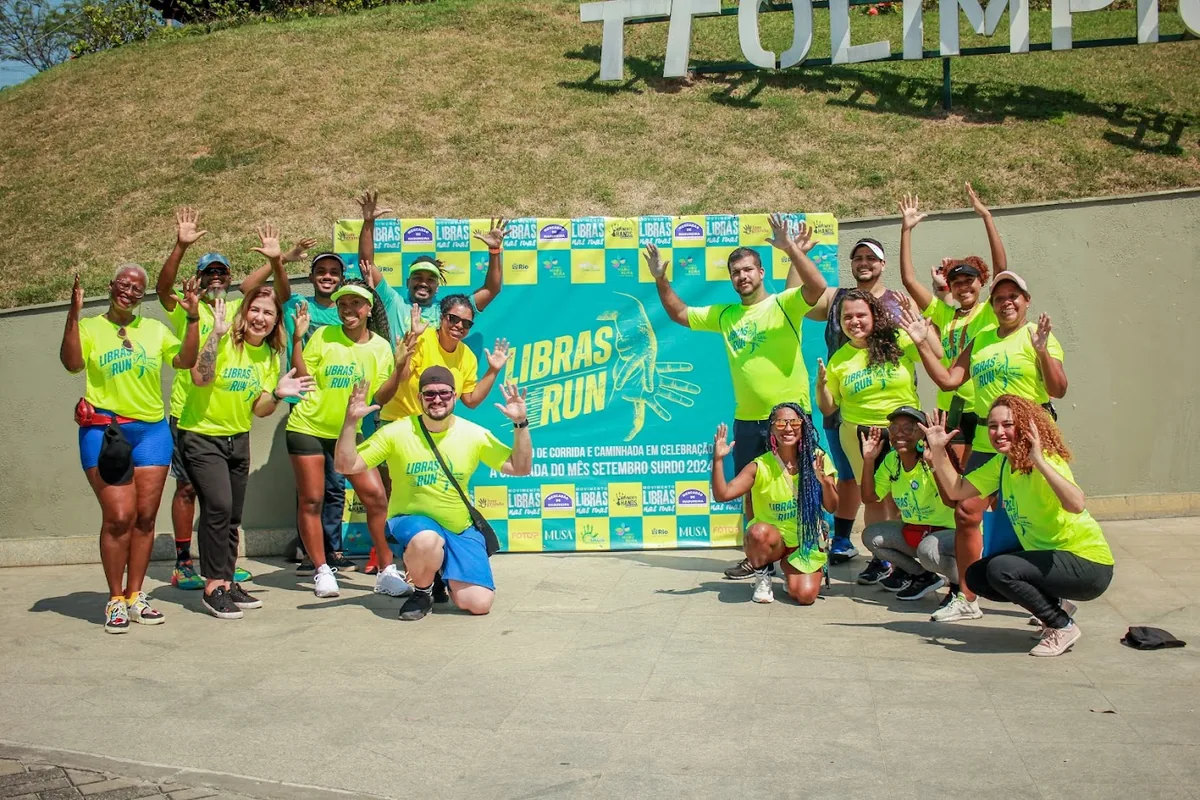 Equipe e participantes do Libras Run de camisetas verdes e amarelas, com os braços para o alto, diante do letreiro Cidade Olímpica