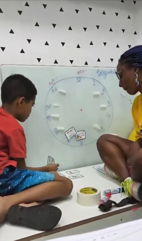 Criança montando um relógio com cartas durante o reforço escolar, ao lado da professora